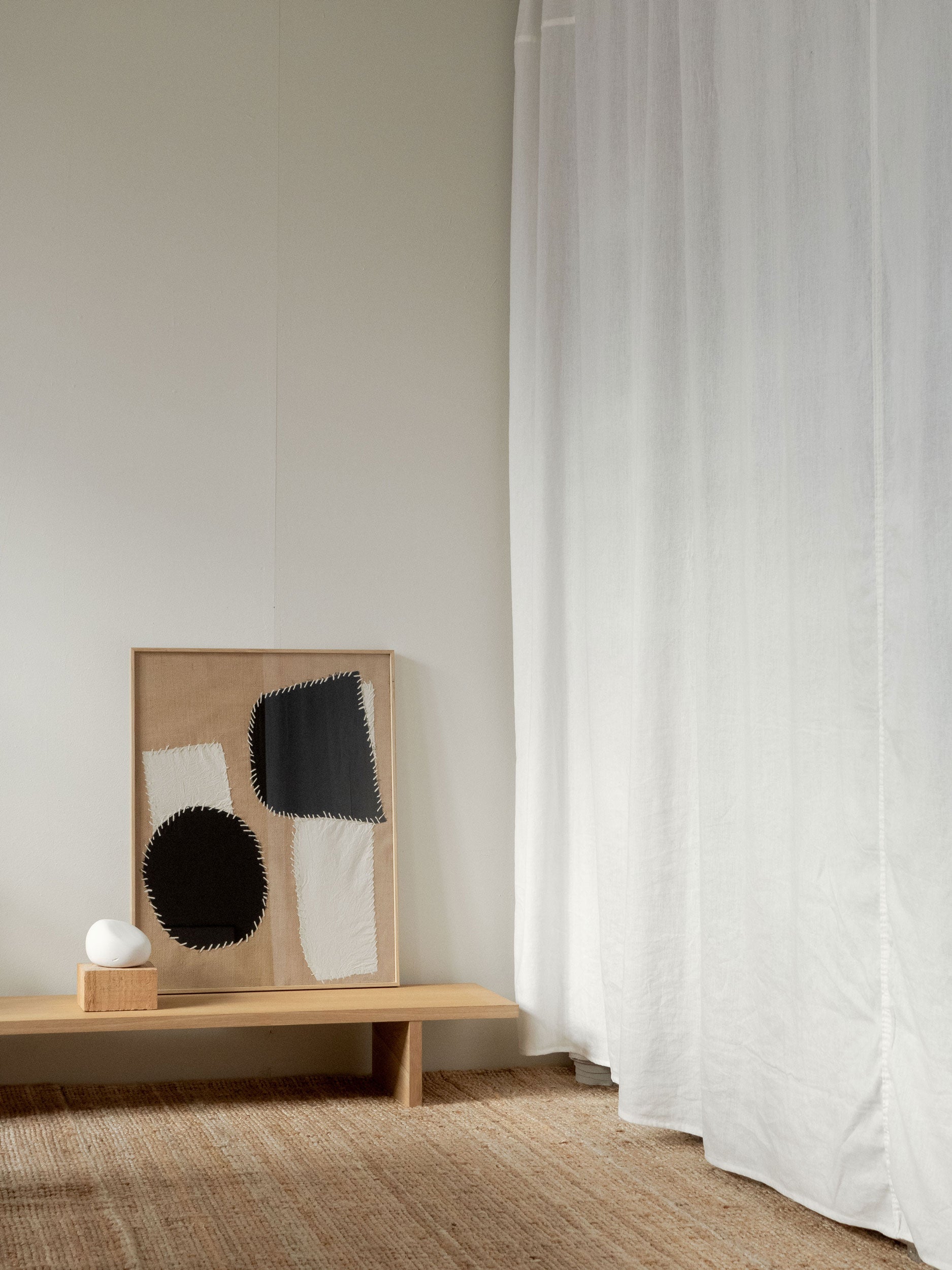 Minimalist room with a wooden bench, and white curtain. On the bench is a minimalist textile artwork with black and white abstract fabric shapes on a hessian background, framed in oak, created as a limited edition by Atelier Cph