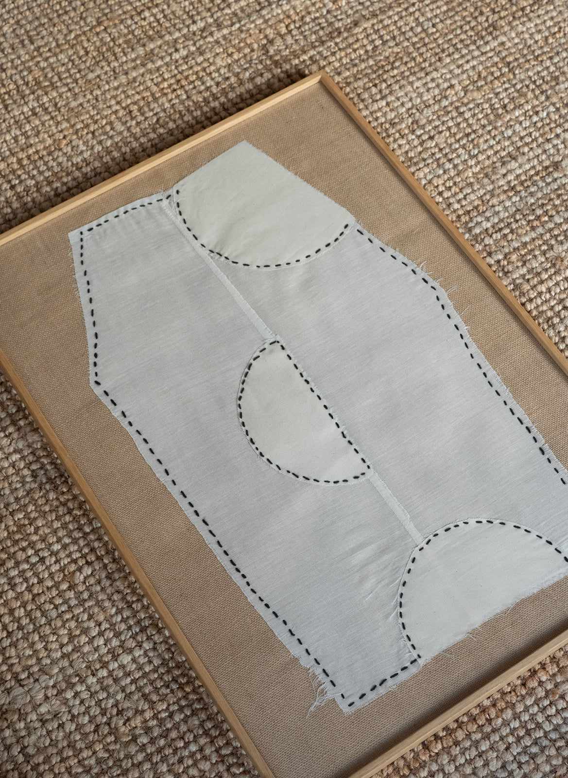 Minimalist textile artwork with white fabric, black stitched lines and curved shapes on a sand-coloured fabric background, framed in oak, created as a limited edition by Atelier Cph.