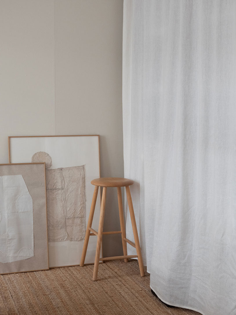 Wooden stool against a wall with framed artwork and a white curtain. Art pieces are made by Atelier Cph