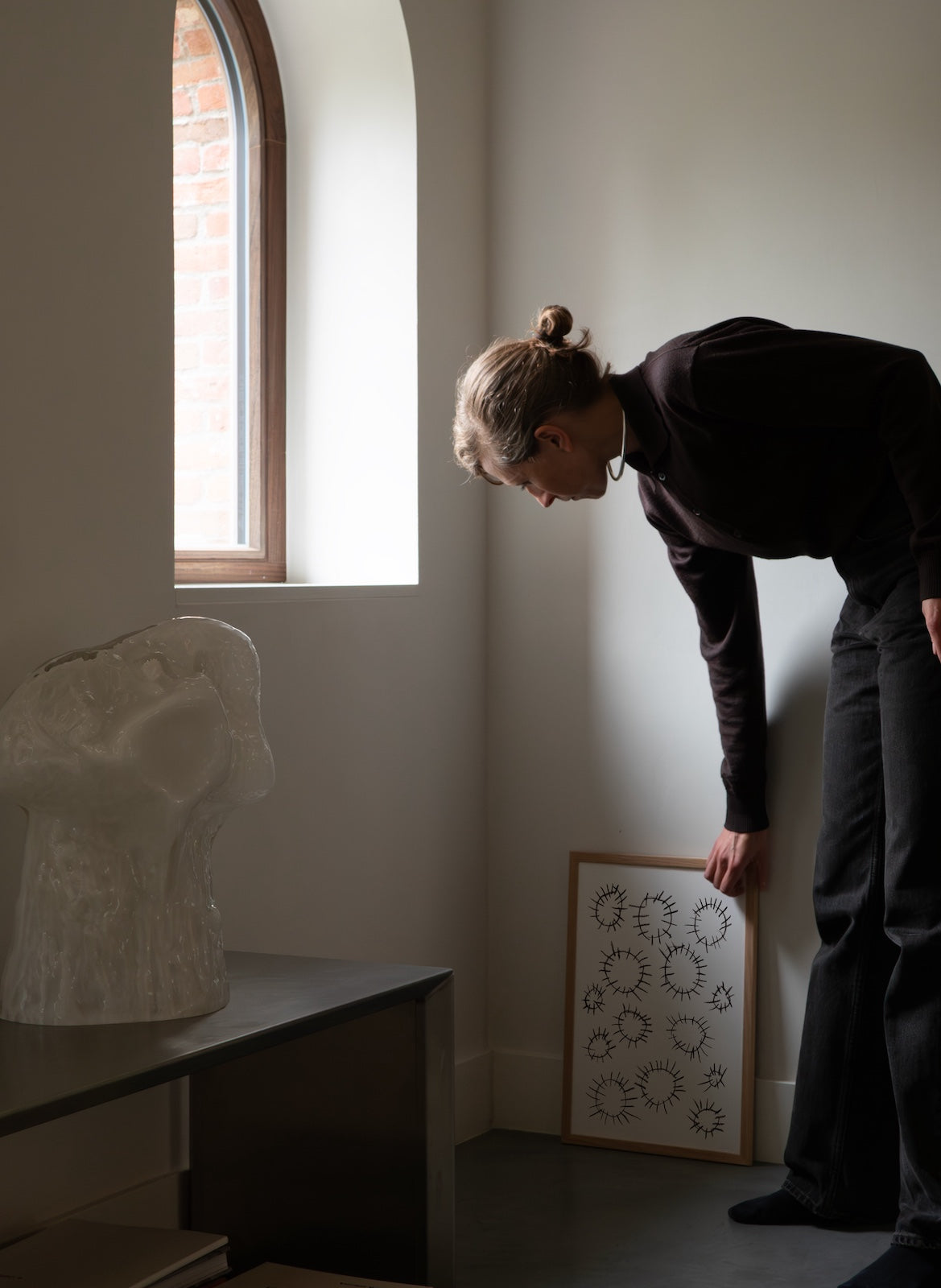 Person observing a sculpture in a room with a window and framed artwork.