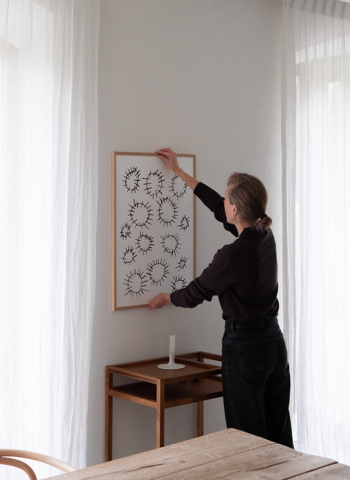 Person hanging a framed artwork on a wall in a room with curtains and a table.