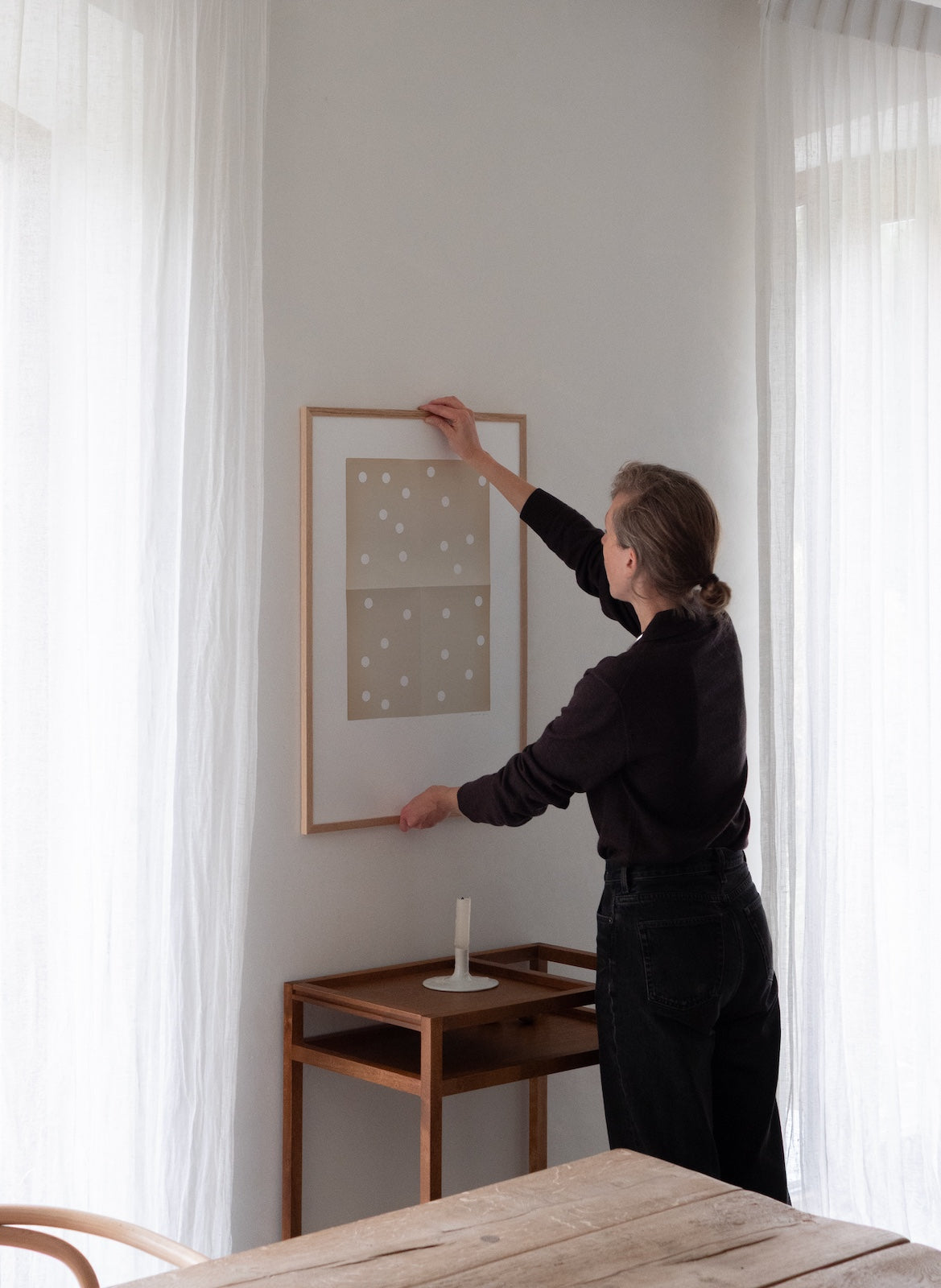 Person hanging a framed picture on a wall in a room with curtains and a wooden table.