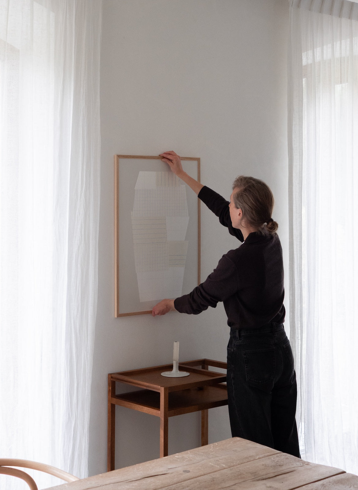 Person hanging a framed artwork on a wall in a room with curtains and a table.