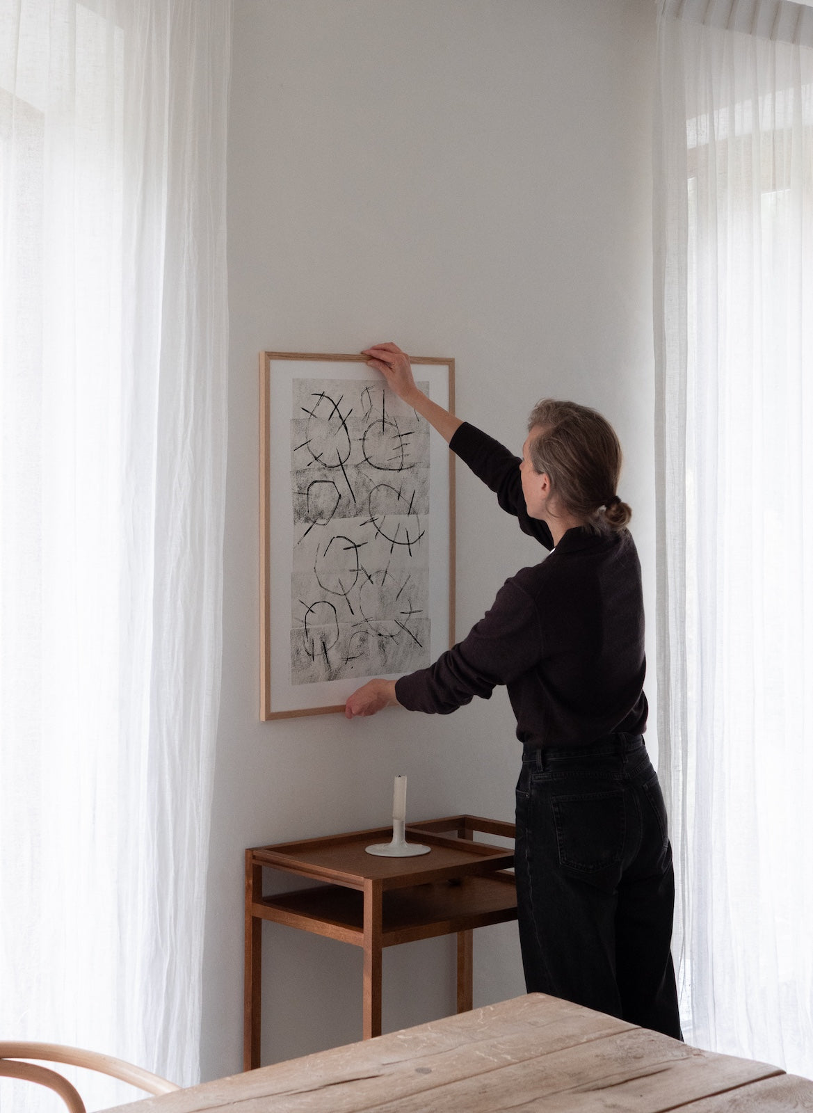Person hanging a framed picture on a wall in a room with curtains and a wooden table.
