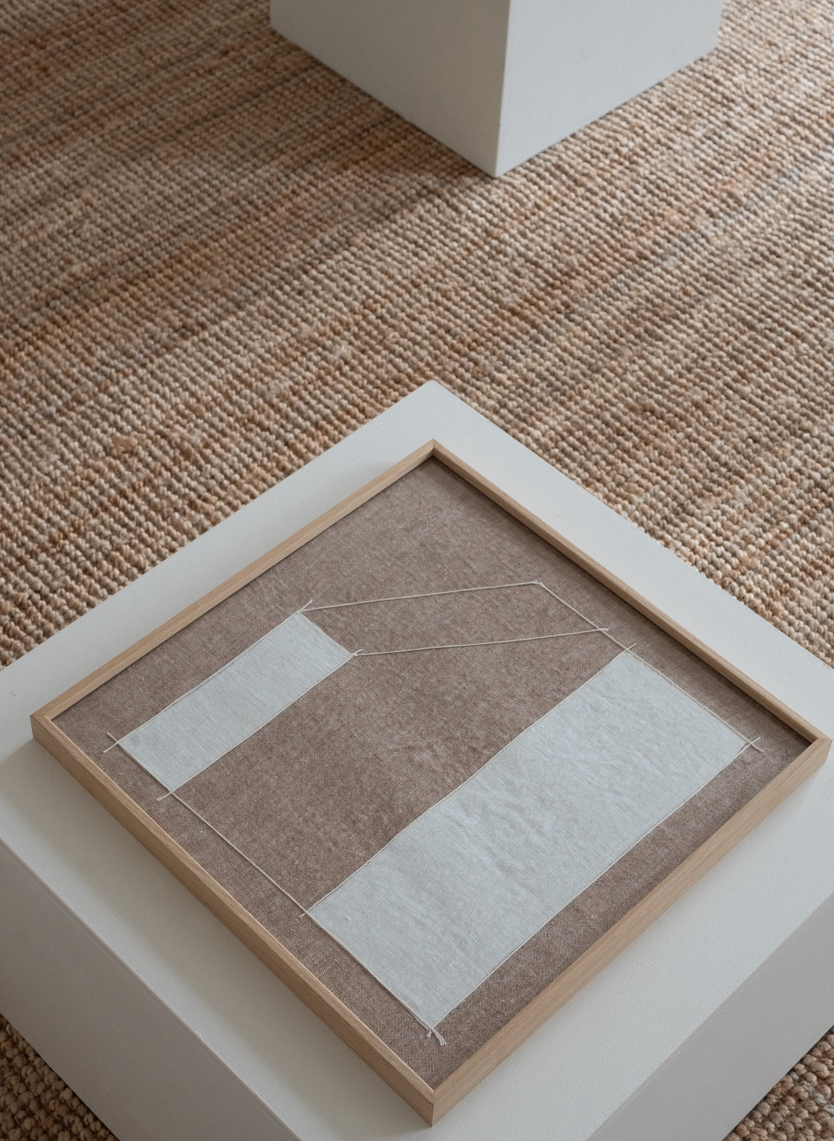 A square framed abstract art print with a light wood frame, featuring a textured taupe background and two white rectangular blocks, displayed on a white pedestal over a woven brown rug.