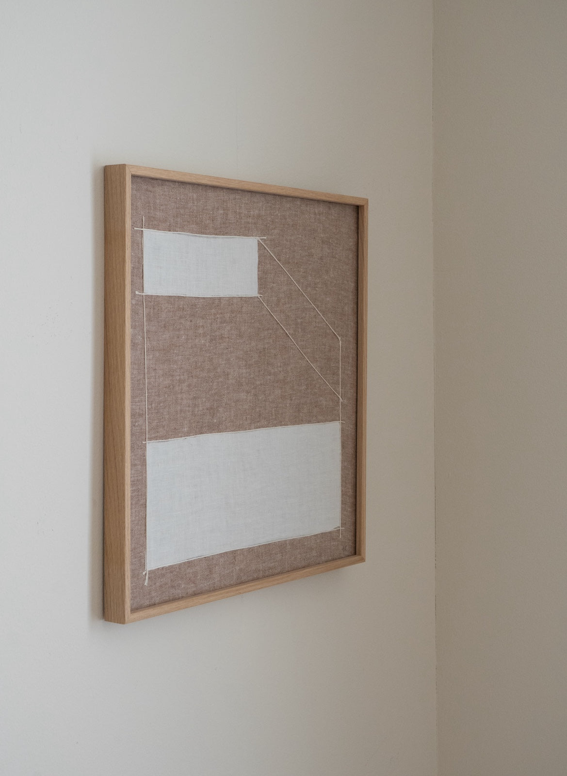 A framed abstract textile wall art piece hanging on a beige wall. The artwork features a light wood frame with a textured taupe linen background and two large white fabric panels.