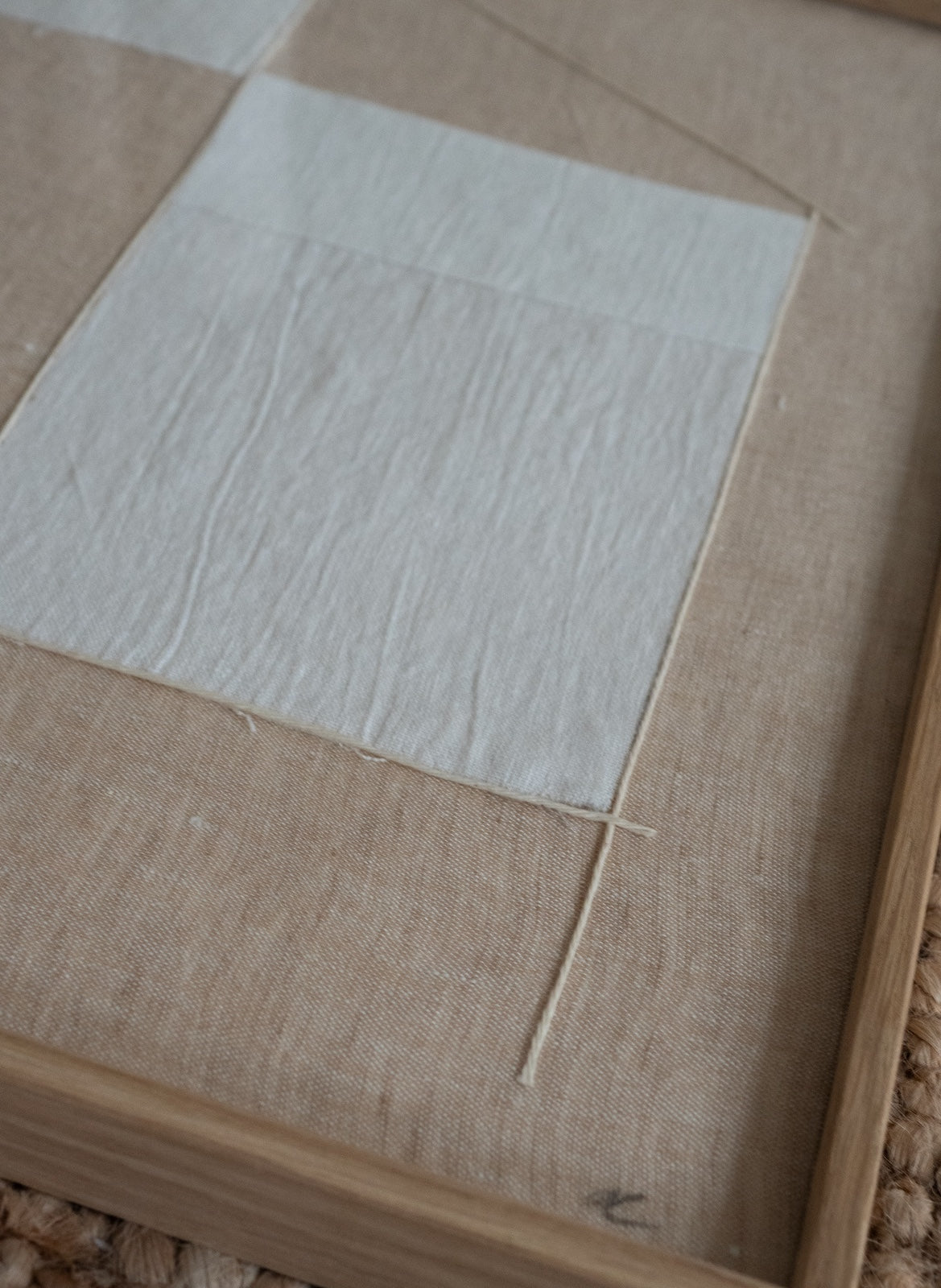 A close-up of a framed textile artwork or box with a light beige linen background, a large off-white square patch, and a natural wood frame.