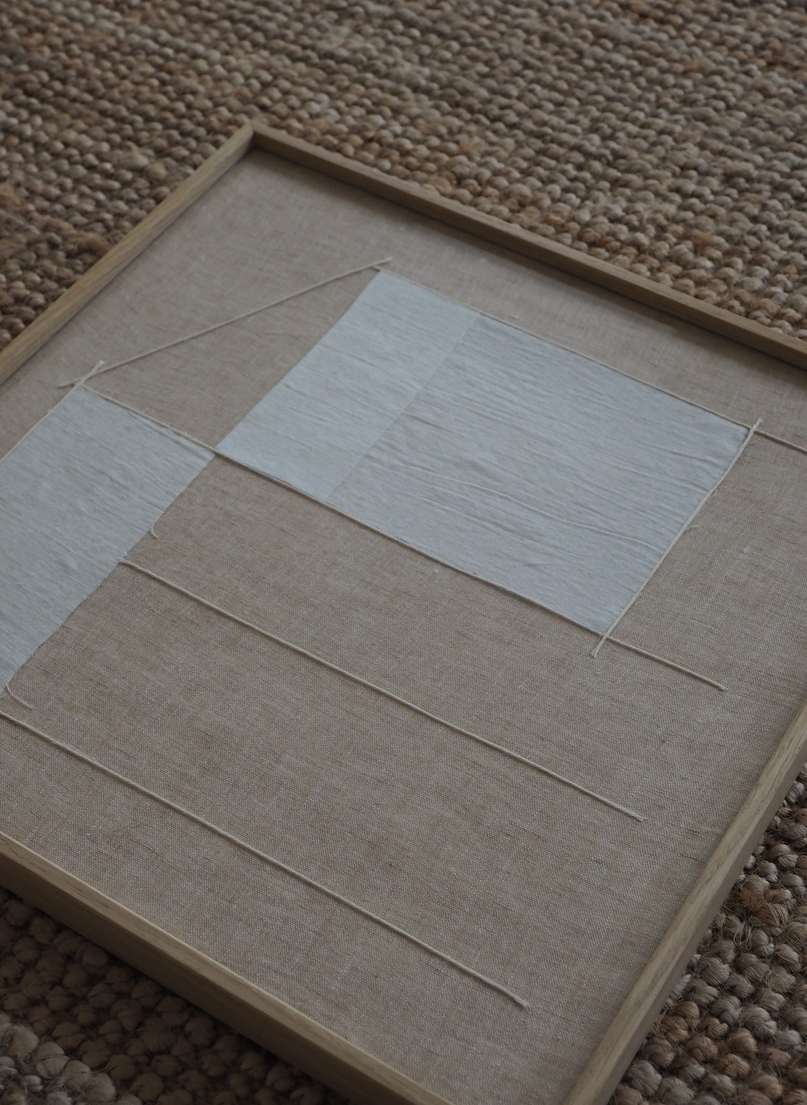 A square framed textile art piece featuring a minimalist geometric design with white fabric panels on a natural linen background, resting on a woven brown rug.