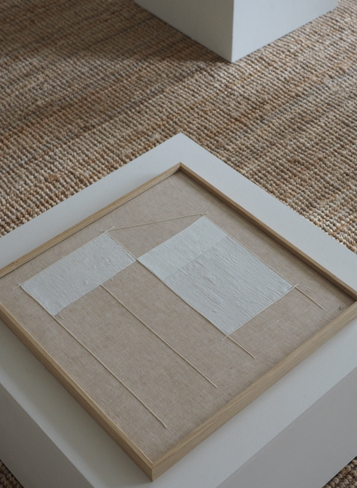 A square framed abstract textile artwork displayed on a white pedestal. The piece features two off-white rectangular fabric panels stitched onto a beige linen-like background with thin stitched lines, set in a light natural wood frame.