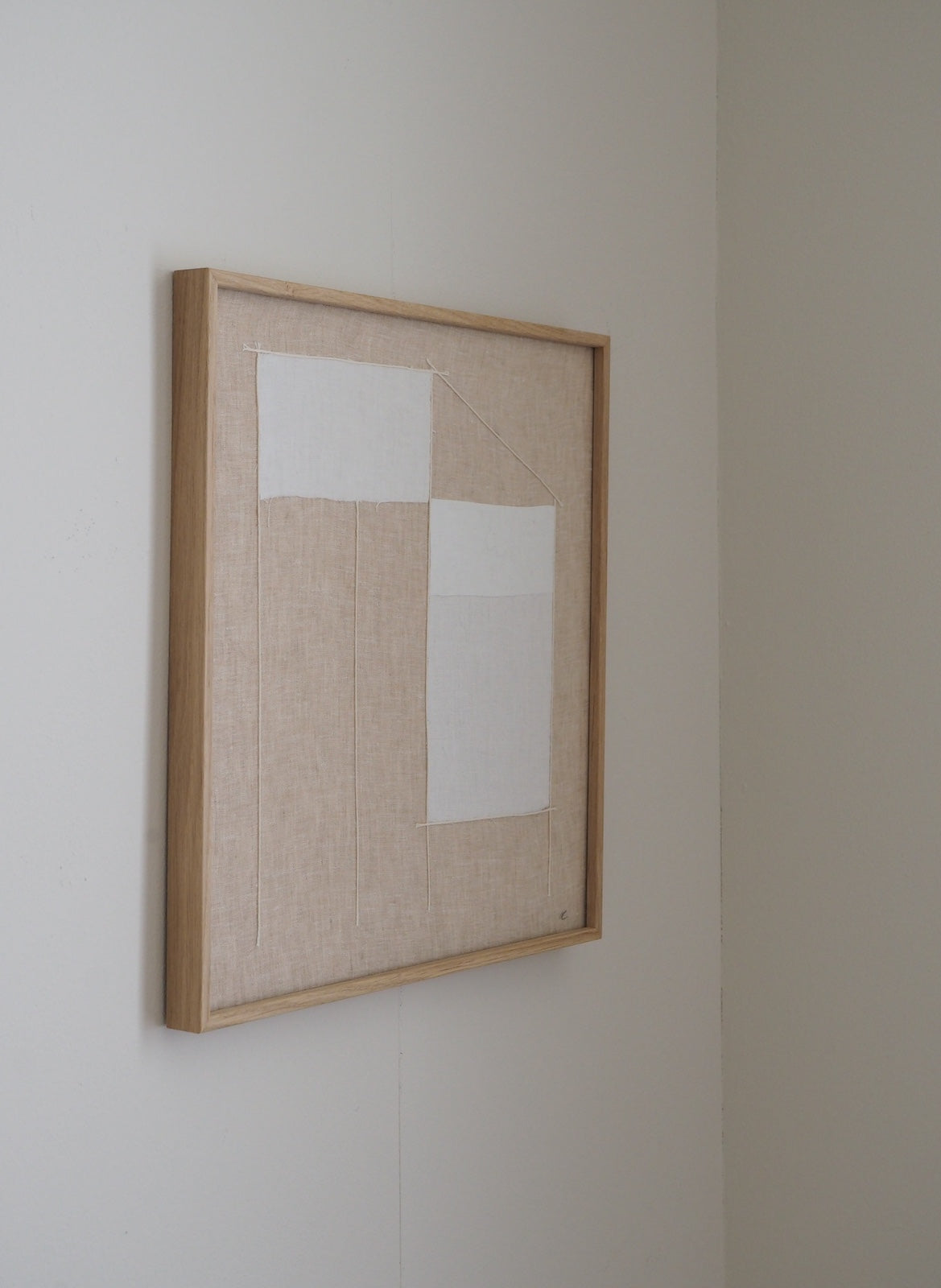 A square framed abstract textile art piece hanging on a light beige wall. The artwork features a light wood frame, a beige linen-like background, and two white rectangular fabric panels.