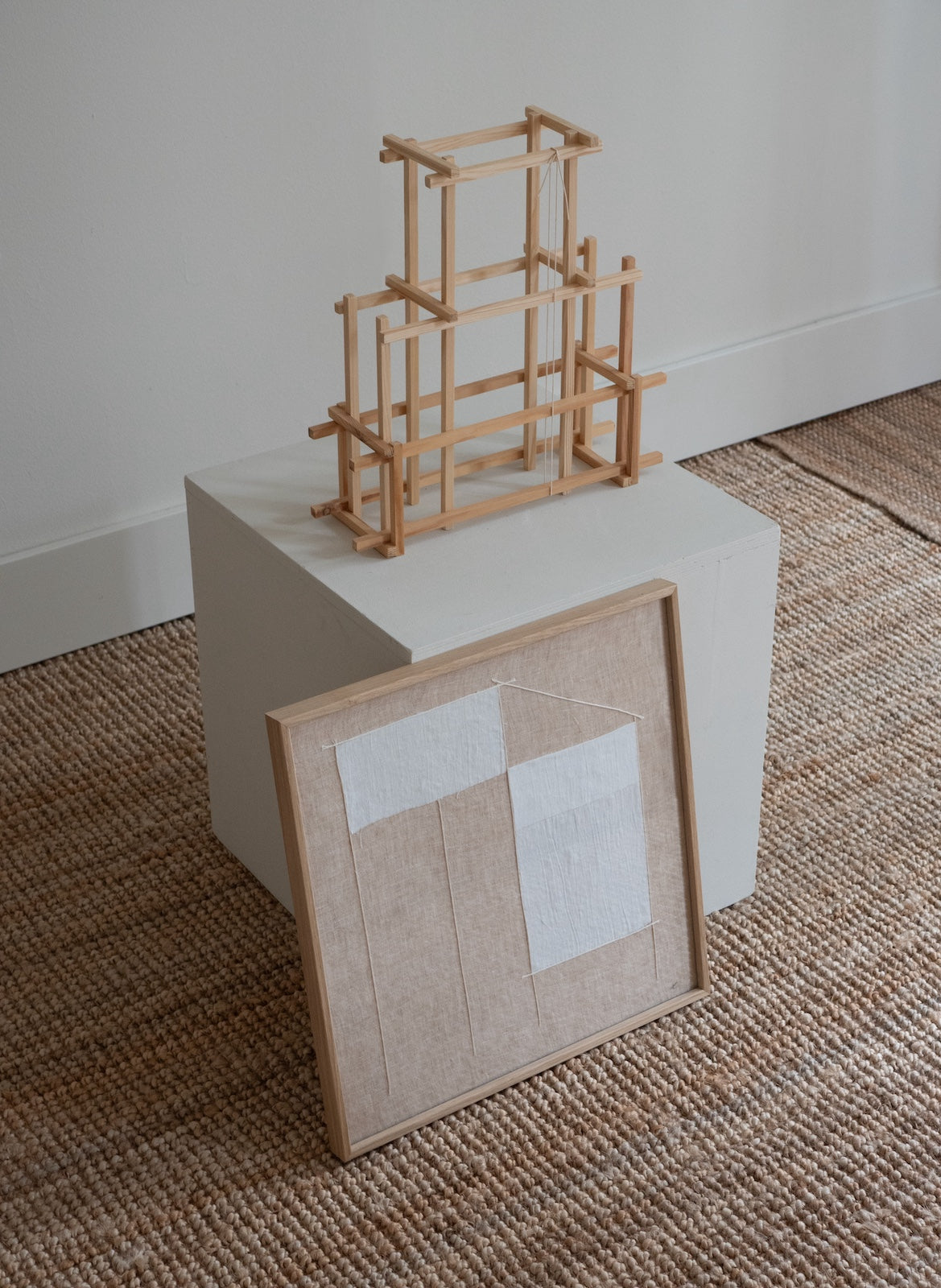 A small wooden geometric frame structure displayed on top of a white cube, with a framed fabric panel leaning in front, set on a woven rug against a plain wall.