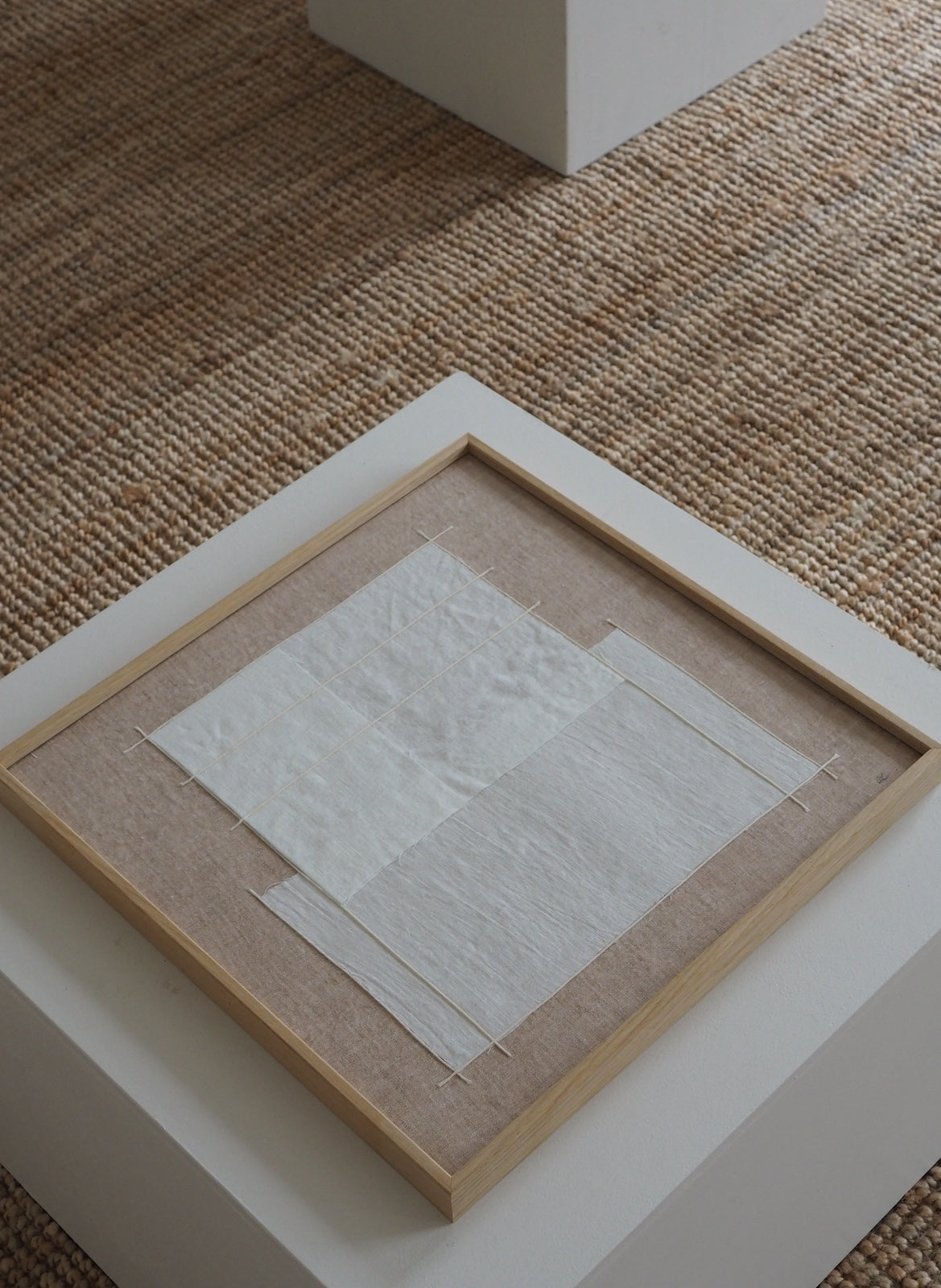 A square framed textile art piece displayed on a white pedestal. The artwork features layered, textured white fabric sheets mounted on a beige linen background, enclosed in a light natural wood frame.