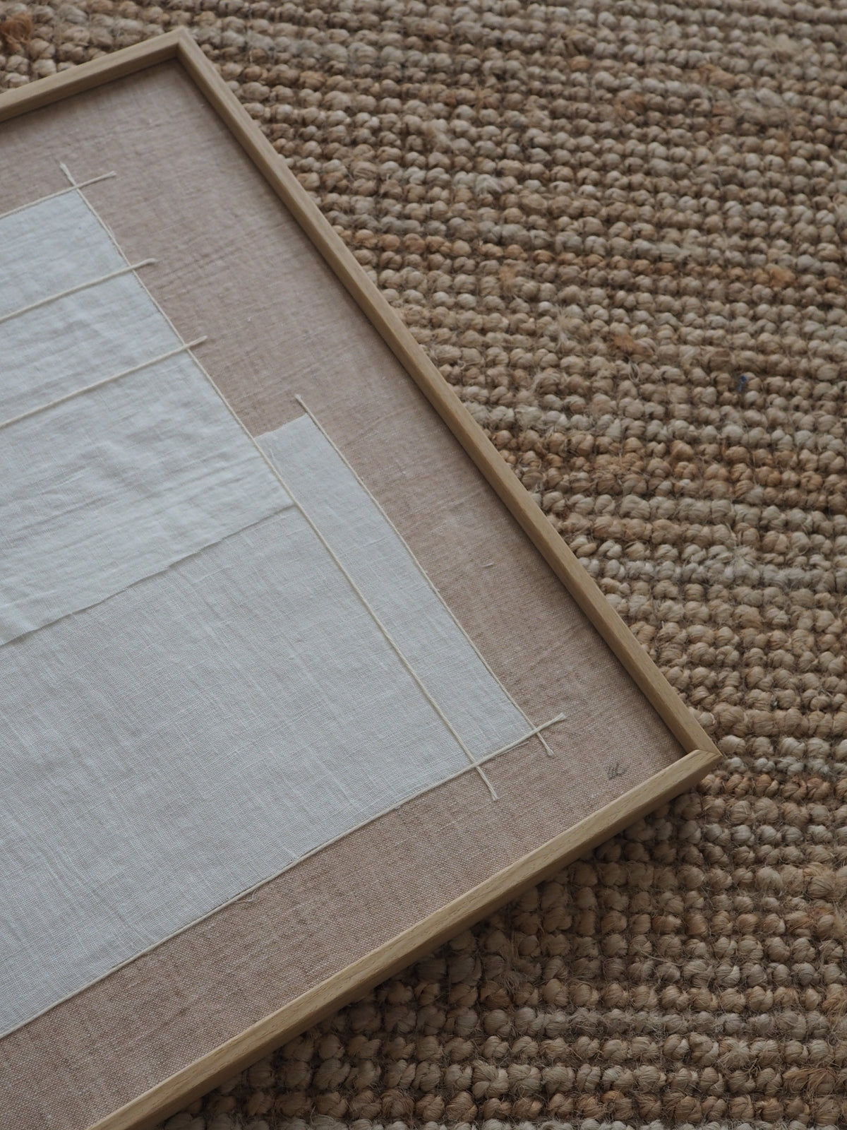 A close-up of a framed textile art piece featuring off-white linen panels mounted on a natural jute background, set on a woven brown rug.
