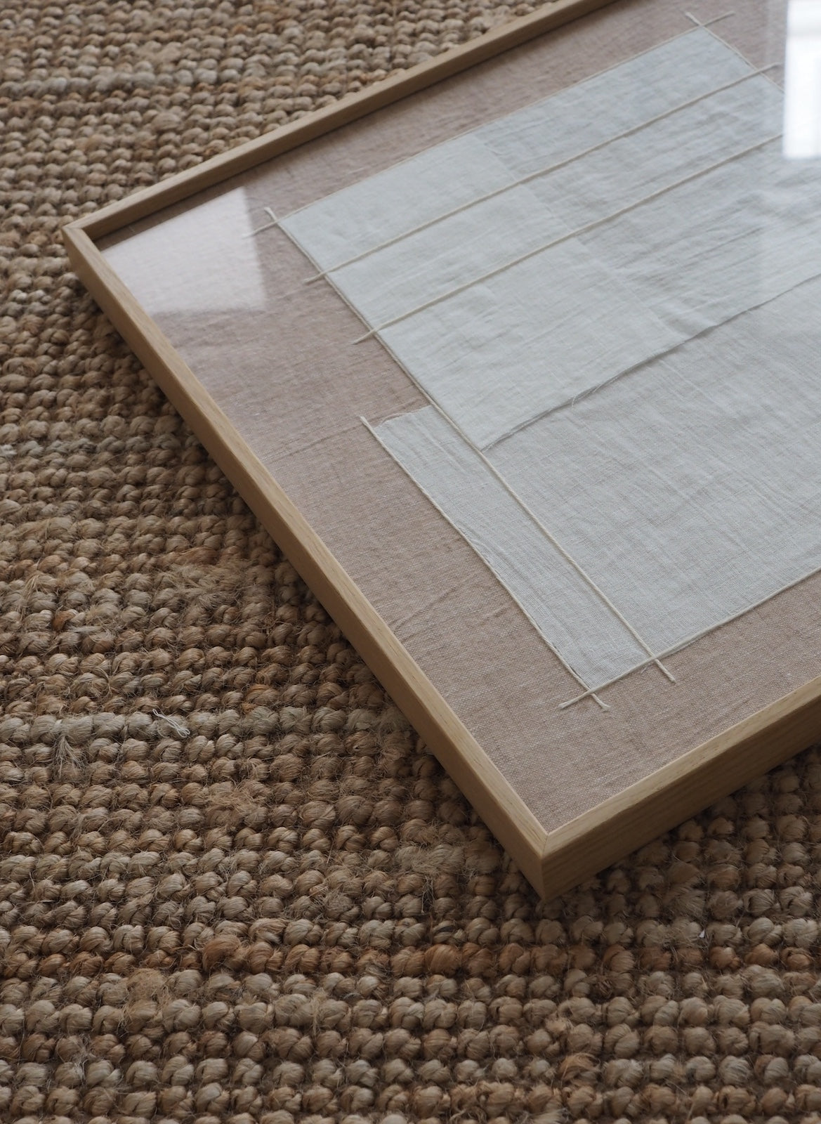 A square framed textile art print with a light wood frame and clear glass, resting on a textured brown woven rug.