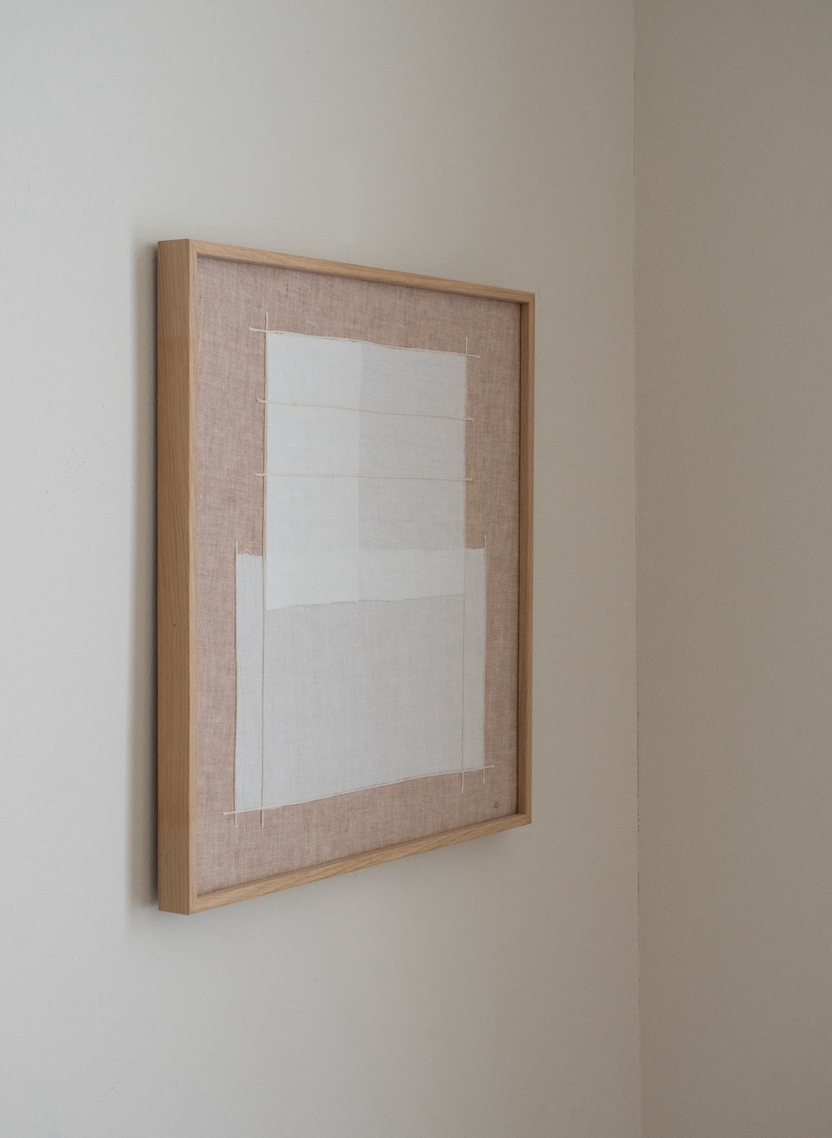 A minimalist framed abstract art print hanging on a neutral beige wall. The artwork features a light wood frame, a textured beige linen border, and a central white geometric grid design.