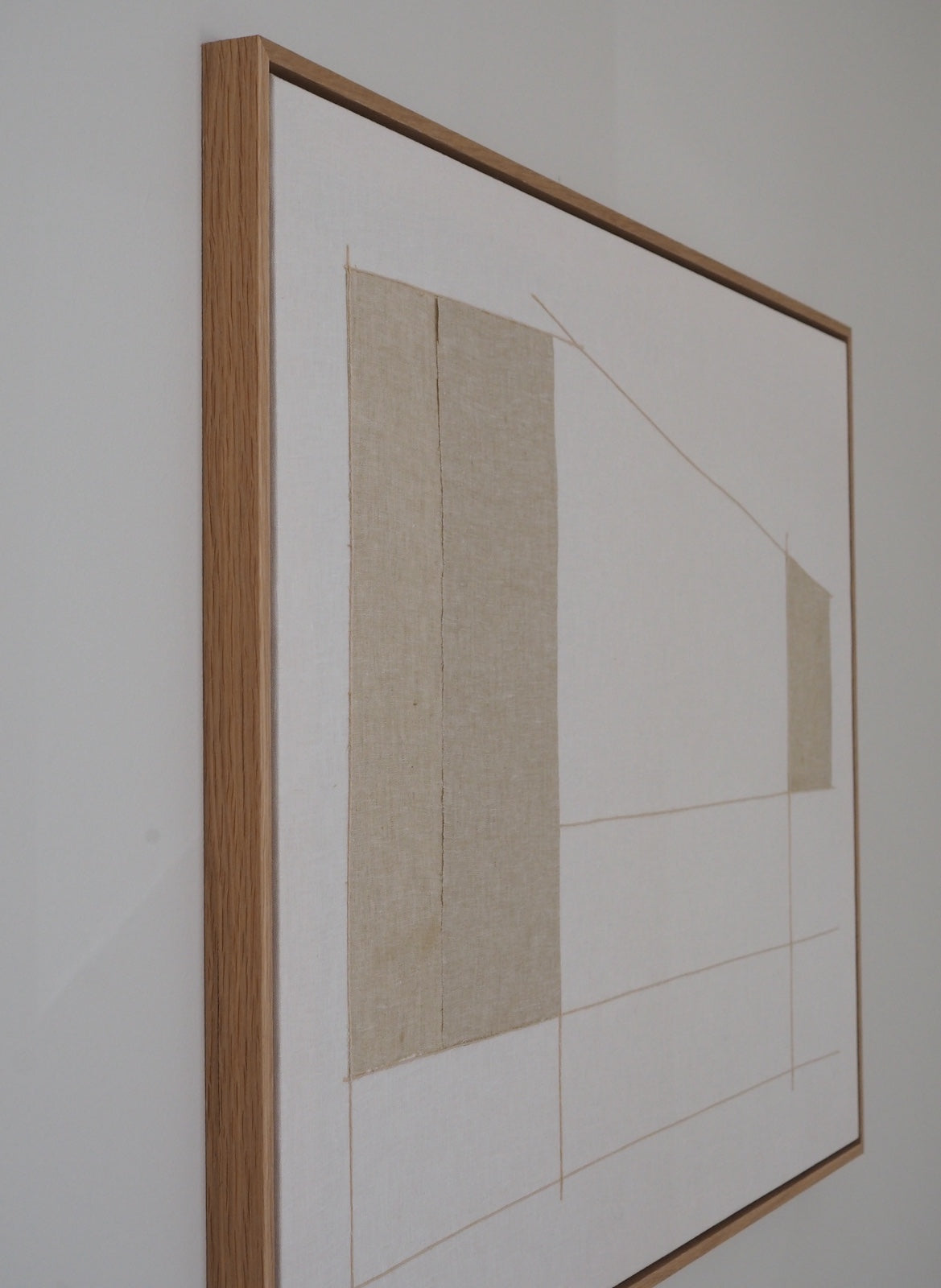 A framed abstract art print featuring minimalist beige geometric shapes and fine lines on a white background, enclosed in a light oak wood frame and hung on a neutral wall.
