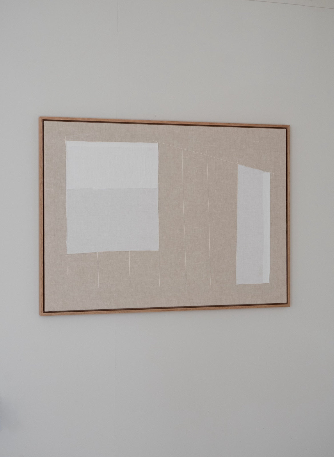 A minimalist framed abstract art print featuring two white geometric shapes on a textured beige linen background, enclosed in a thin light brown wooden frame and mounted on a neutral gray wall.