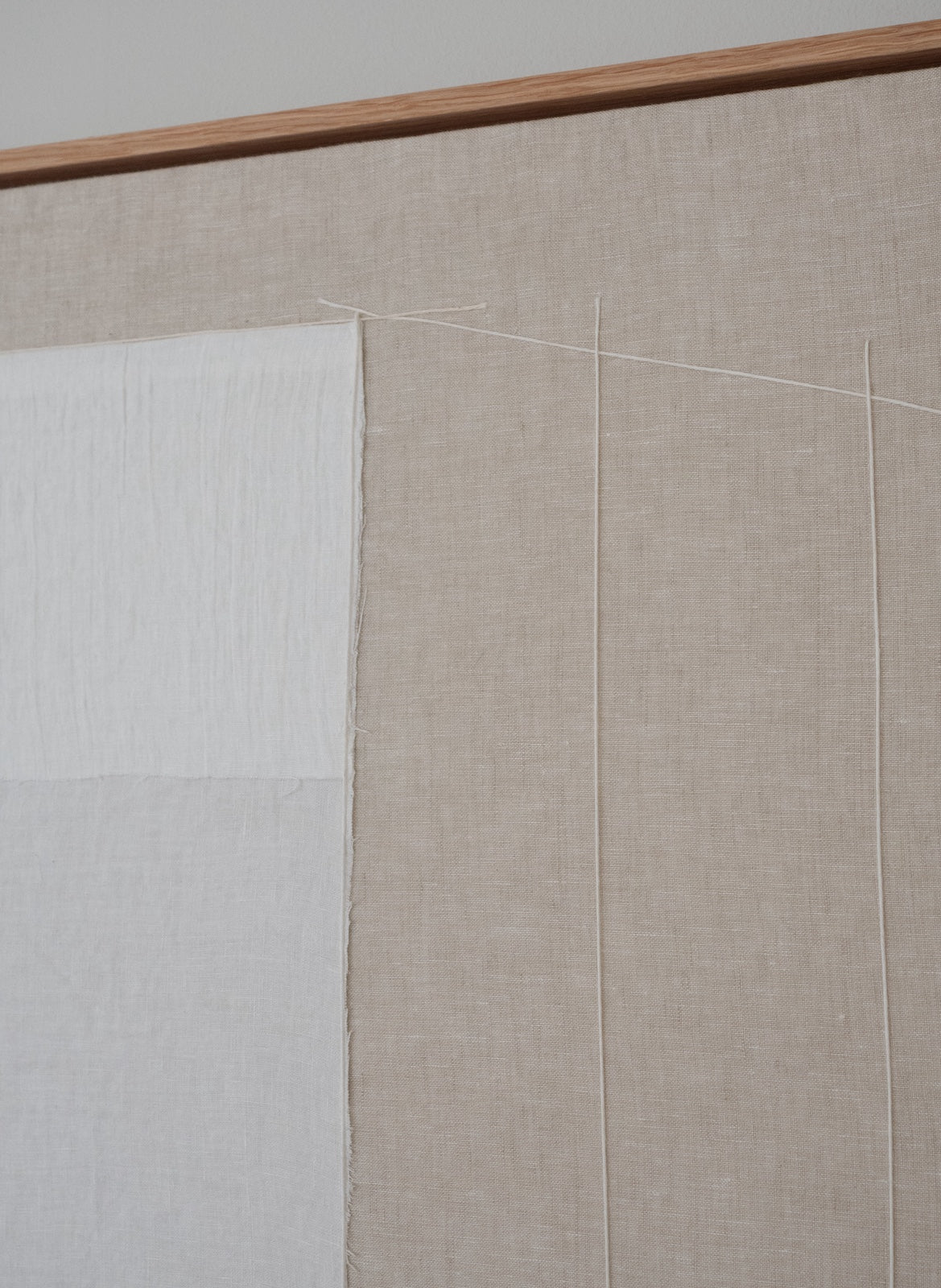 A close-up of a textile wall hanging or fabric panel with a natural beige linen-like background, a large white fabric panel on the left, thin vertical stitched lines, and a wooden rod along the top edge.