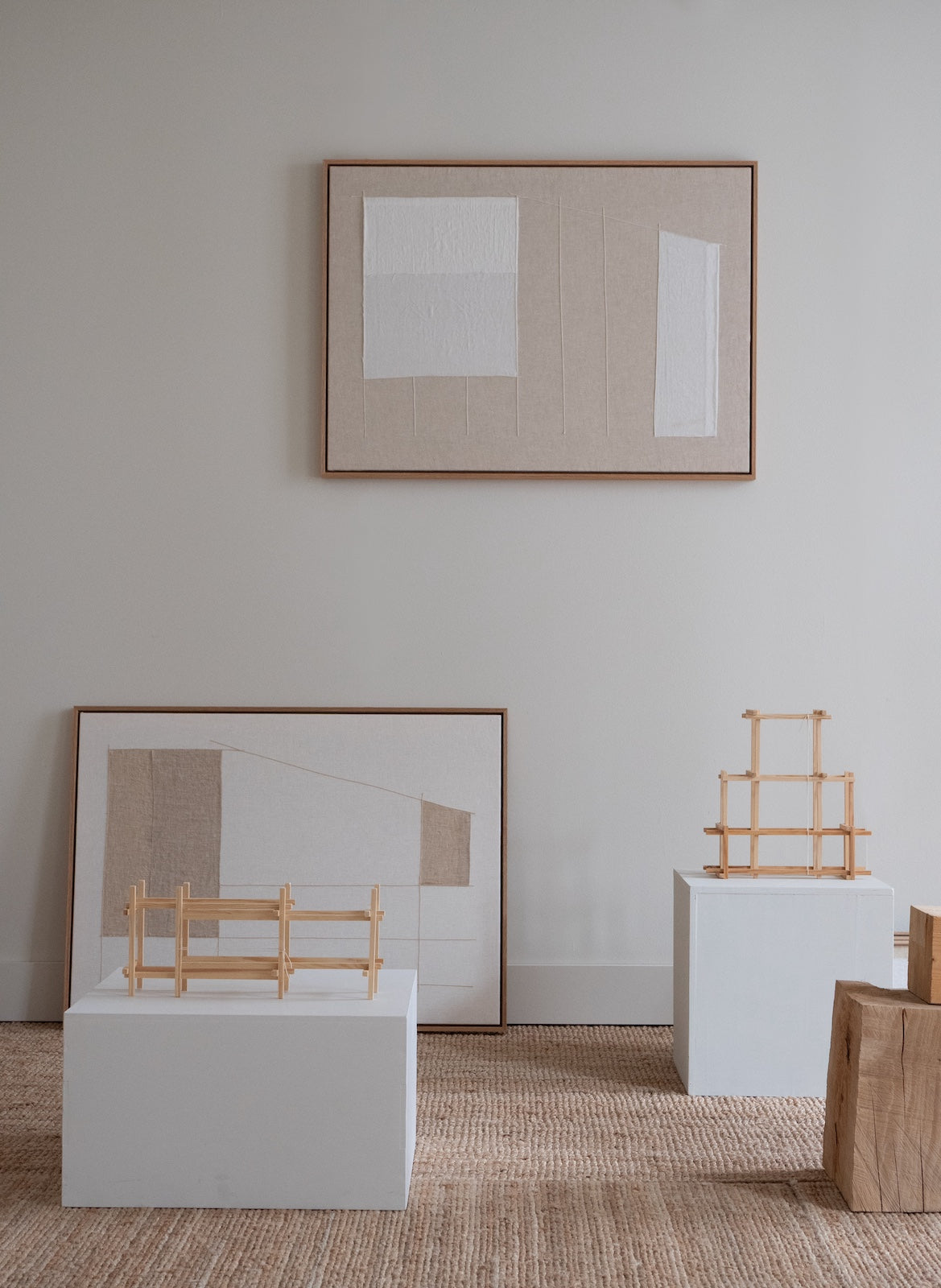A minimalist interior scene with three framed abstract artworks in thin wood frames, displayed on white pedestals against a light gray wall. The artworks feature neutral beige, white, and tan geometric shapes.