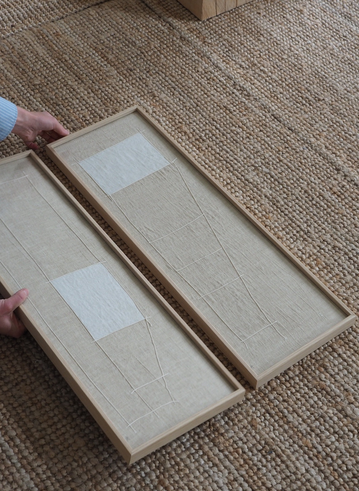 Two rectangular framed wall art pieces with beige linen-like panels and white rectangular patches, held by hands on a woven jute rug.