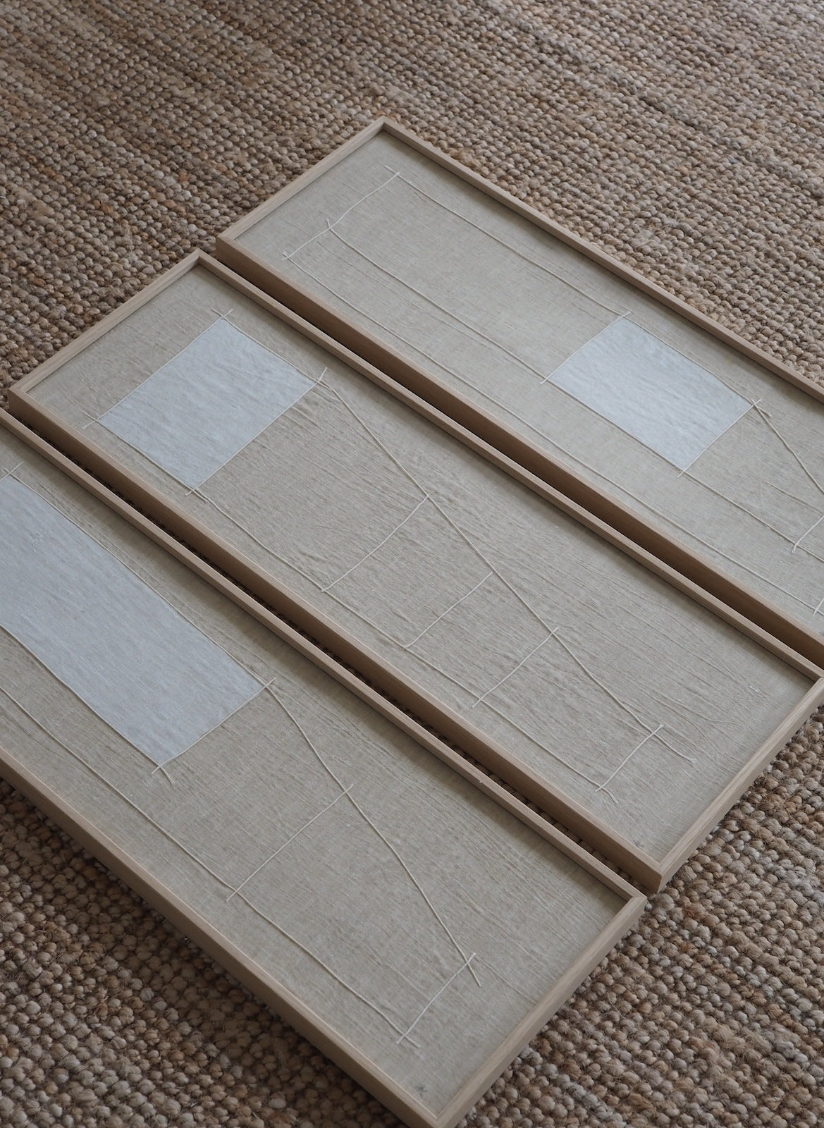 Three rectangular framed art panels with textured beige linen-like surfaces and white rectangular patches, arranged diagonally on a woven brown rug.