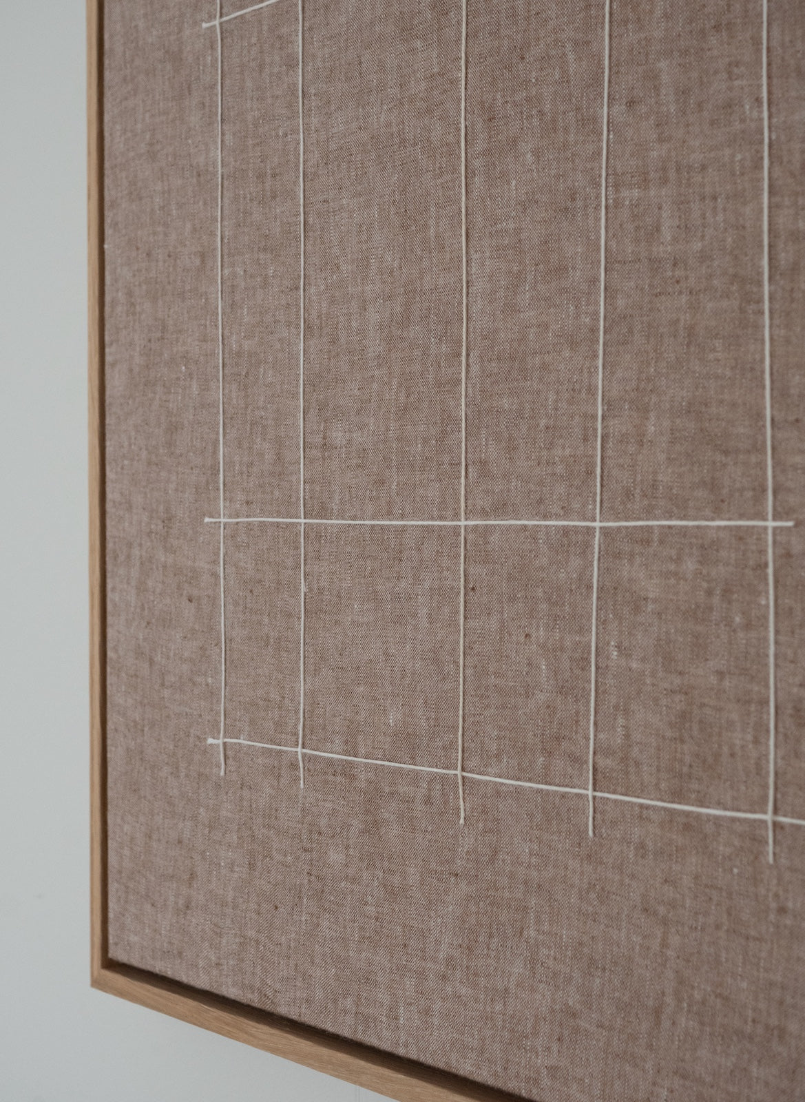 A close-up of a linen fabric board in a warm taupe-brown color, decorated with a white grid pattern and framed in a light natural wood.