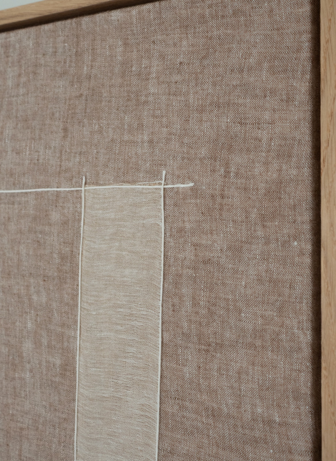 A close-up of a woven fabric surface in warm taupe-brown with a vertical strip of lighter beige fabric and thin white thread details.