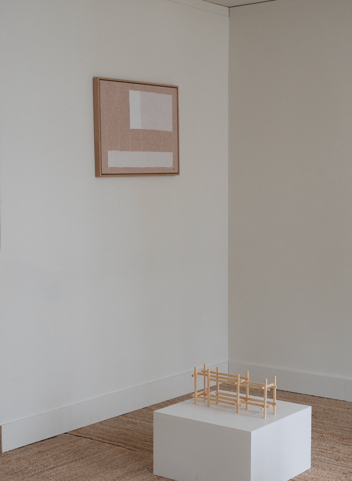 A minimalist framed abstract art print hangs on a light beige wall in a modern room, with a small wooden geometric model displayed on a white pedestal in the foreground.
