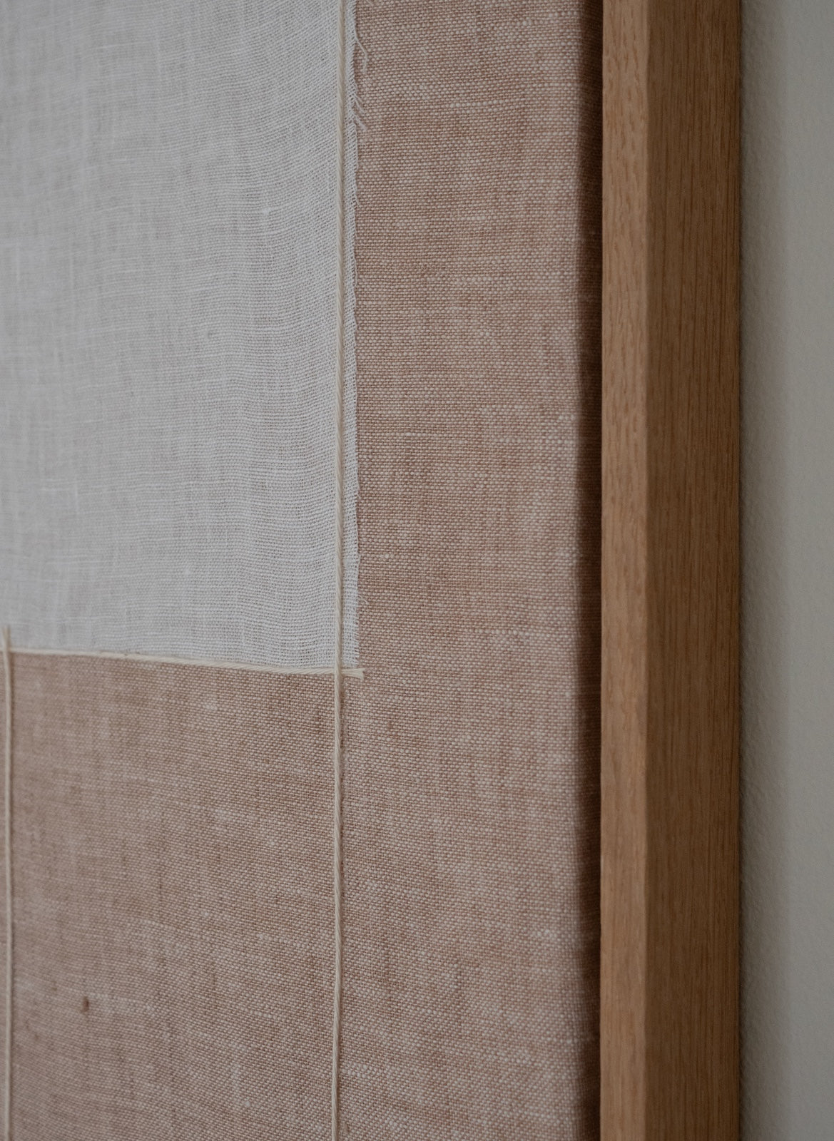 Close-up of a framed textile wall art piece with two linen panels in off-white and warm beige, bordered by a natural wood frame on the right.