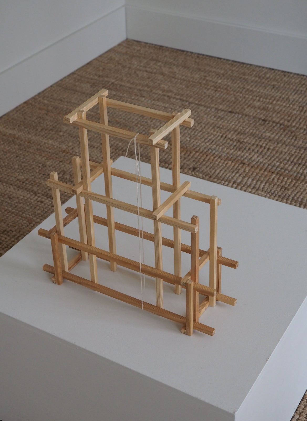 A minimalist wooden geometric sculpture or frame structure displayed on a white pedestal in an indoor setting with a woven rug and white walls.