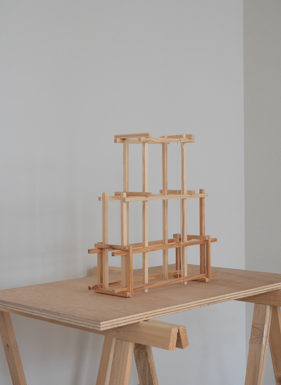 A small wooden frame structure made of light natural wood stands on a plywood table against a plain light gray wall.